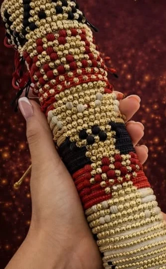Mano sosteniendo un rollo de pulseras bambalinas tejidas en rojo, negro y crema con balines de oro.