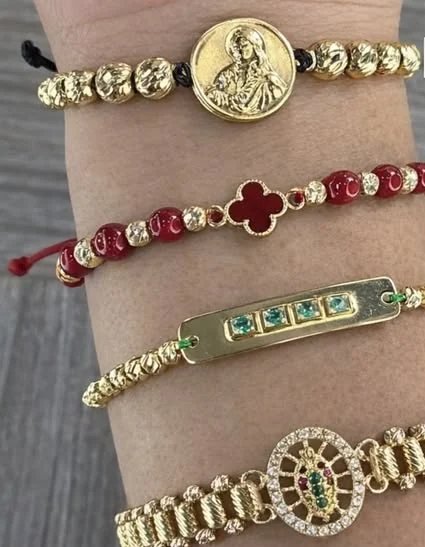 Cuatro pulseras doradas en un antebrazo: Sagrado Corazón, trébol rojo, barra con esmeraldas y rueda con circonitas.