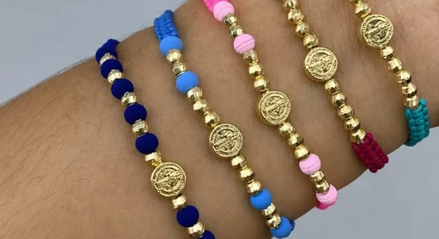 Pulseras tejidas de colores con medalla de San Benito en oro, puestas en una muñeca.
